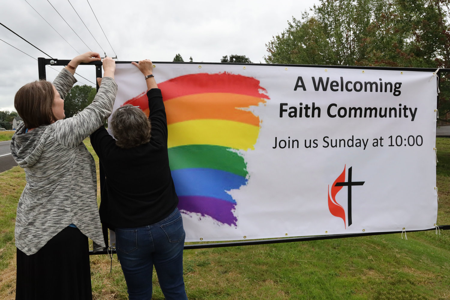 From left, Battle Ground Community United Methodist Church member Kristin Wade and pastor Susan Boegli hang up a banner in front of the church July 17. The banner is a replacement from a prior one with the same message on it that Boegli said was stolen recently, an act she felt was motivated by the banner’s intent of supporting LGBT individuals. 